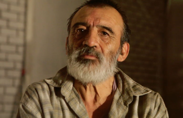 A headshot photograph of a man wearing a check shirt. The man has a beard is looking at the camera.
