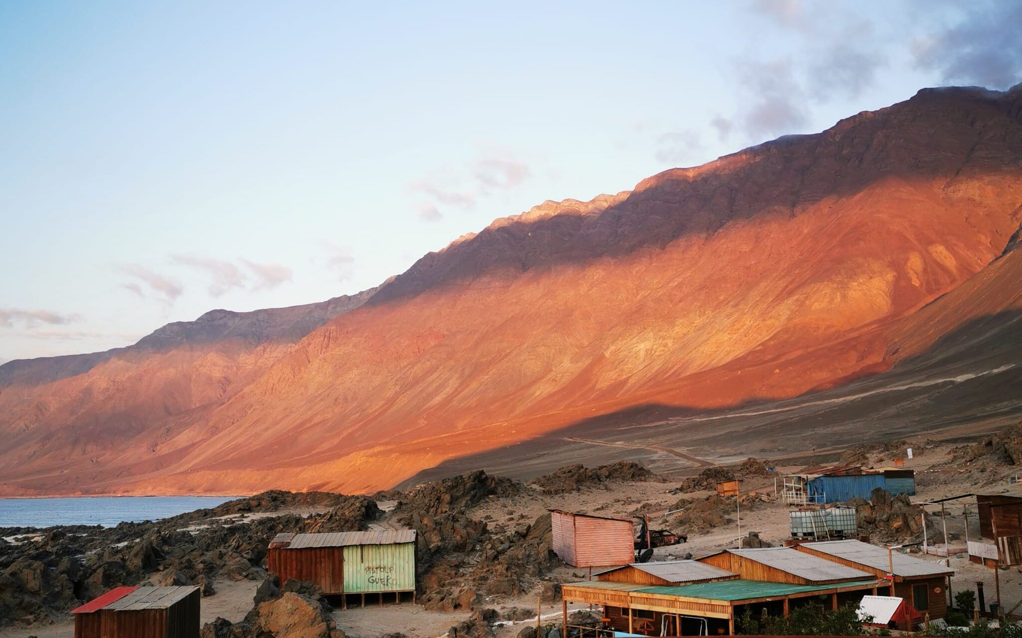 There is a red cliff face beneath a blue sky that stands against a blue shore line. There are shacks in the foreground. The sun is setting.